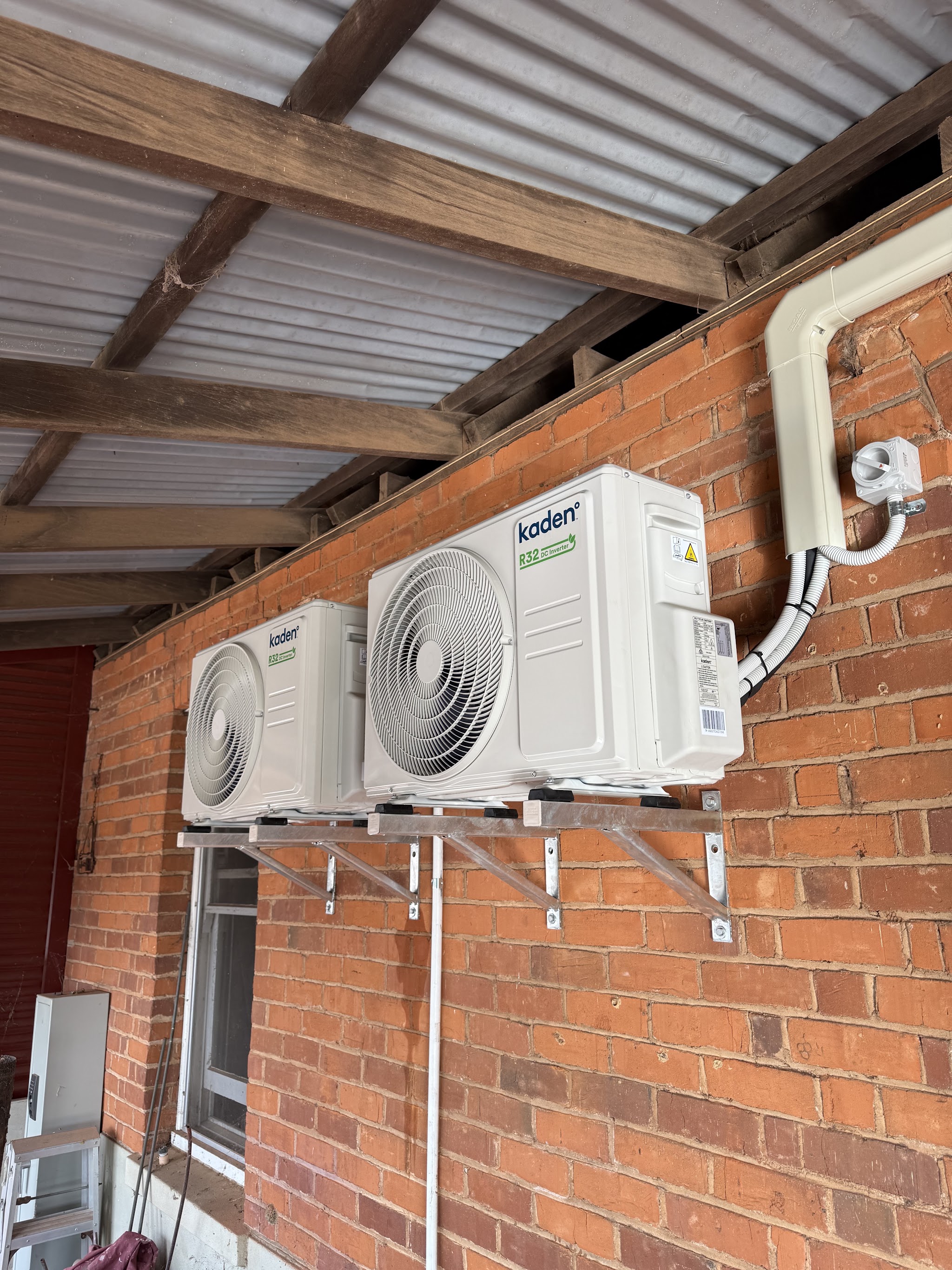 Two Kaden inverter units bracketed high on a red brick wall under a verandah
